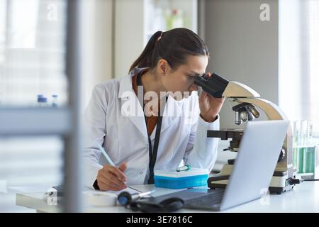 Young adult Caucasian woman scientist examining sample through microscope and taking notes in laboratory. Ideal for medical research, biotech innovation, diagnostics marketing Stock Photo