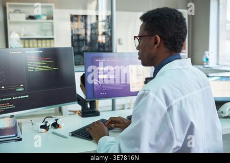 Black male scientist analyzing medical data on desktop monitors, typing in laboratory workspace. Ideal for healthcare technology, diagnostics, research, or AI innovation marketing Stock Photo