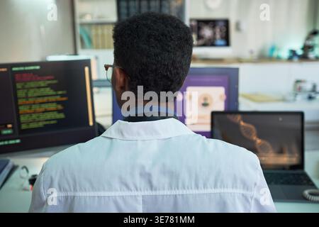 Rear view of young adult Black man scientist analyzing medical data and DNA visuals on multiple screens in lab. Ideal for healthcare technology, research, diagnostics marketing Stock Photo
