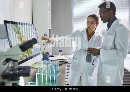 Caucasian young adult woman and Black young adult man scientists reviewing lab data on monitor and discussing research results. Useful for healthcare innovation, biotech teamwork marketing Stock Photo