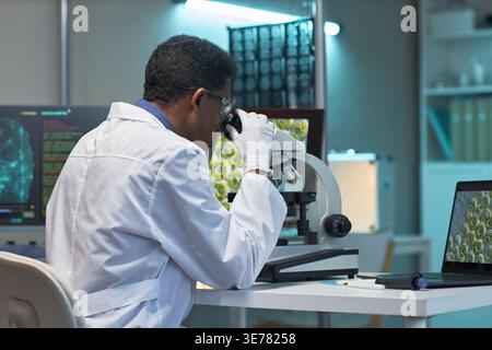 African American male scientist examining specimen through microscope in laboratory, analyzing medical data on screens. Useful for healthcare research, diagnostics, biotech marketing Stock Photo
