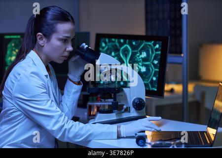 Young adult Caucasian woman scientist examining sample through microscope and using laptop in laboratory. Ideal for medical research, biotechnology, diagnostics, innovation marketing Stock Photo