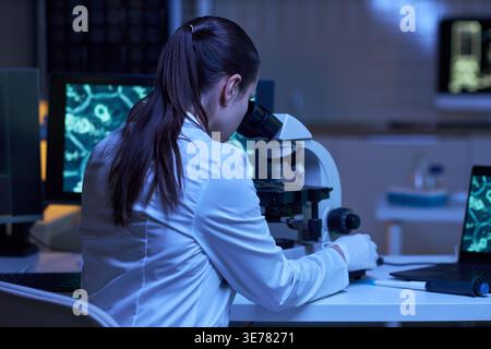 Young adult woman scientist examining sample through microscope in laboratory, supporting medical research and diagnostics. Ideal for healthcare innovation, biotech, testing campaigns Stock Photo