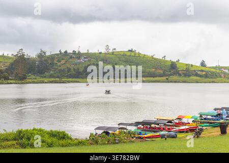 A serene lake environment with green hills, small boats docked along the shore, and a motorboat creating ripples on the water under a cloudy sky. Nuwa Stock Photo