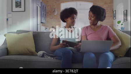 African American mother and teen daughter sitting on gray sofa at home, sharing tablet and laptop Stock Photo