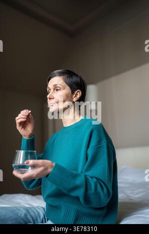 Senior woman taking special medicine to cure illness, holding glass of water and pill supplement. Stock Photo