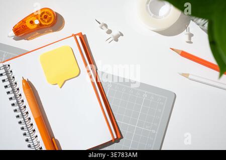 Open notebook with sticky note, orange correction tape, and cutting mat on a white desk. Copyspace Stock Photo