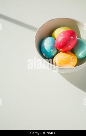 Five bright colorful Easter eggs in bowl on white table with copy space. Festive homemade Easter eggs under bright morning sunlight. Easter traditions Stock Photo