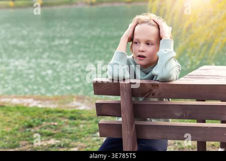 Thoughtful boy resting wooden bench calm lake autumn park hold head hands looking away across water and yellow foliage. Quiet outdoor moment shows Stock Photo