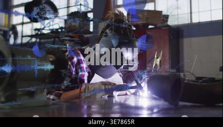 Welding woman welder leaning over bench at shop, in plaid shirt, helmet, holding torch, overlays Stock Photo