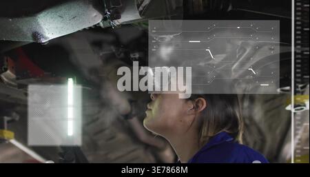 Inspecting female technician in blue coverall examining lifted vehicle chassis at garage, LED HUD Stock Photo