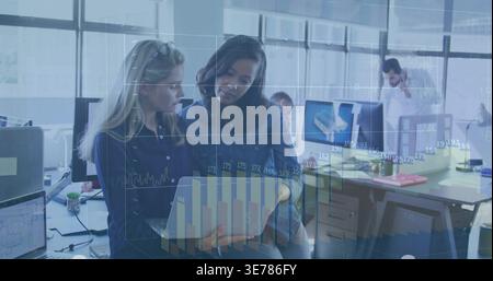Collaborating two women reviewing charts on laptop in open office, wearing denim jacket and blouse Stock Photo