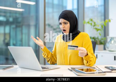Muslim woman in hijab using a laptop at her desk. Holding a credit card and reacting with a surprised and disappointed facial expression while encount Stock Photo