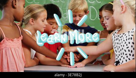 Stacking hands, 7 pupils joining at school table wearing casualwear, chalkboard Learn More overlay Stock Photo