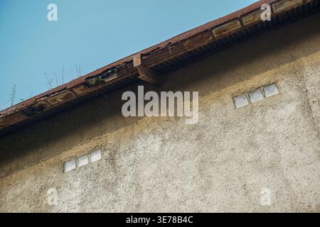 The rough textured plaster of the building's walls with small checkered vents and a corrugated iron roof that looks rusty and badly moss Stock Photo
