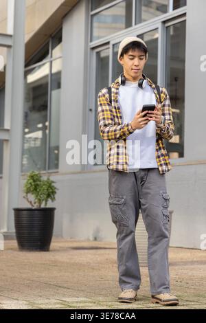 Asian man standing on sidewalk by windows wearing plaid shirt using phone with backpack Stock Photo