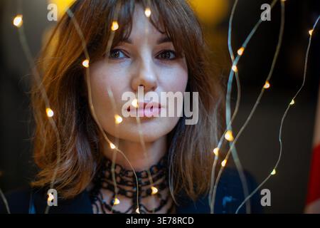 Woman with brown hair stands in front of decorative lights, creating a warm atmosphere that accentuates her features and stylish outfit Stock Photo
