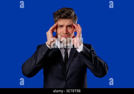 30s aged businessman in formal wear having headache, studio portrait. Guy putting hands on head, isolated on blue background. Concept of problems, medicine, illness Stock Photo