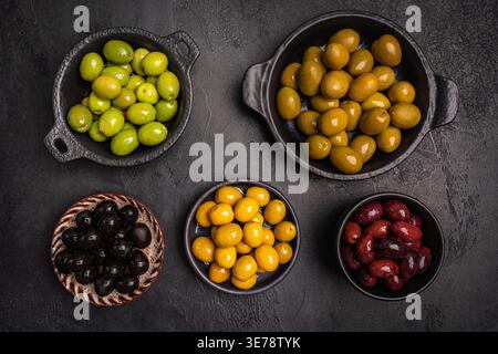 Green and black olives in a bowls and olive oil on dark background top view. Olive oil and variety of green and black olives in bowls with olive leaves all around taken from above. Stock Photo