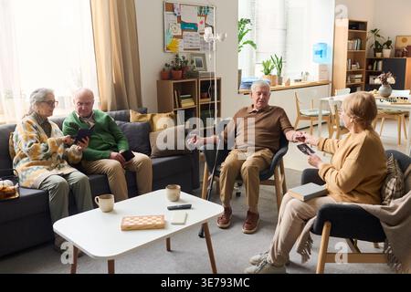 Senior men and women socializing in care home lounge, sharing photos and using smartphone during group activity. Useful for retirement living, elder care, community support marketing Stock Photo