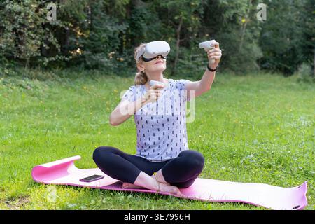 A mature woman smiles while interacting with a virtual environment using VR goggles and controllers. This image highlights active aging, technological Stock Photo