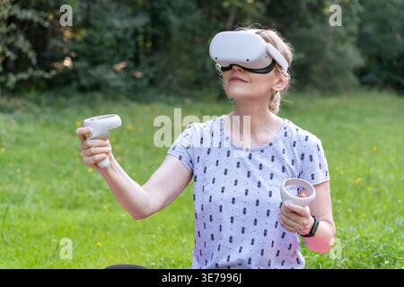 A focused woman smiles slightly while interacting with a virtual environment using VR goggles and controllers in a park. This image highlights technol Stock Photo