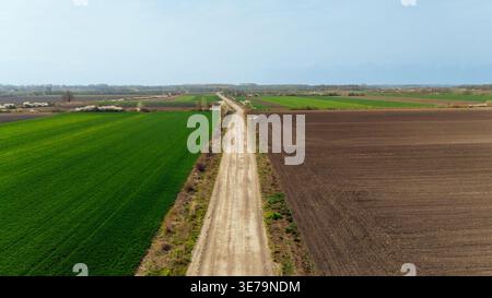 Aerial view of a long dirt road slicing through vibrant green fields and freshly plowed brown earth, contrasting textures under a soft sky, Sremska Mi Stock Photo