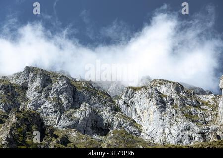 View of craggy, gray mountains rise, their peaks shrouded in ethereal white clouds against a backdrop of deep blue sky, Picos de Europa, Asturias, Spa Stock Photo