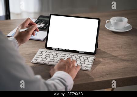 Hands using tablet with blank screen mockup and keyboard on desk with calculator document and coffee cup, performing typing and data entry workflow fo Stock Photo