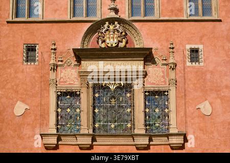 Ornate Renaissance window framed by carved stone, decorative ironwork and gilded coat of arms on a warm terracotta Prague facade; architectural detail Stock Photo