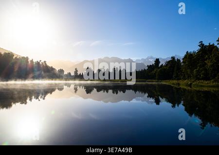 View of a serene lake mirroring the snow-capped mountains under a radiant sun, with mist rising gently over the still water, Lake Matheson, New Zealan Stock Photo