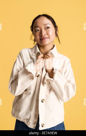 A confident young Asian woman stands stylishly against a bright yellow wall. Stock Photo