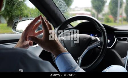 Young businessman experience to ride an autonomous self driving electric car at rainy day. Male businessperson resting during riding on electrical Stock Photo