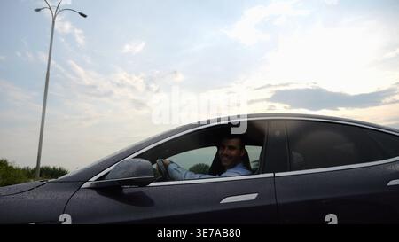 Young businessman driving on electric car in circular path on asphalt road after rain. Happy manager showing grimaces and fooling around while riding Stock Photo