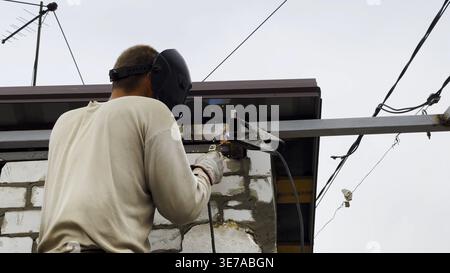 Manual laborer in protective mask welds iron detail at construction. Professional young repairman with welding machine working at the site. Male Stock Photo