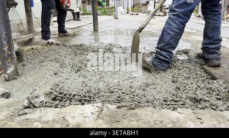 Male hands in protective gloves uses trowel to spread and level fresh cement in yard. Another worker distributes and moves the mixture with a shovel Stock Photo