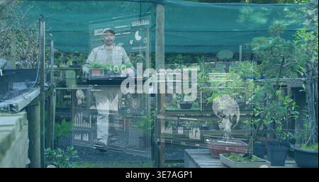 Mature man carrying black seedling tray in light jacket and cap, viewing AR overlays in nursery Stock Photo
