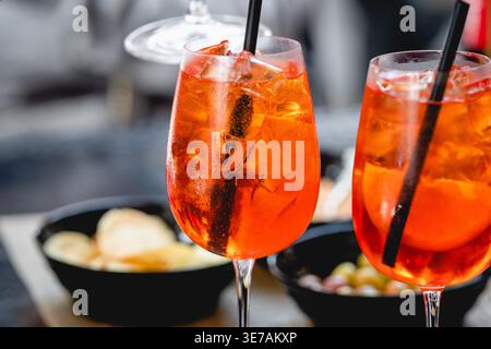 Vibrant orange Spritz cocktails with ice and citrus slices served in wine glasses with straws, accompanied by snacks in a casual dining setting Stock Photo