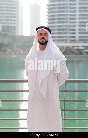 Arab businessman stands in Dubai Marina, showing traditional clothing against a backdrop of modern architecture and water. Stock Photo