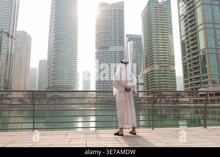 Arab businessman in kandura stands by the water in Dubai Marina looking at the tall buildings. The scene shows modern urban life in UAE. Stock Photo