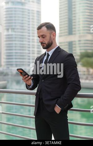 Businessman in formal suit stands by railing using smartphone with Dubai Marina skyline in the background during daytime. Stock Photo