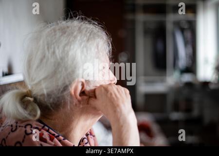 Profile of an elderly woman with white hair looking into the distance indoors. Senior retirement lifestyle, aging at home, thoughtful elderly woman. Stock Photo