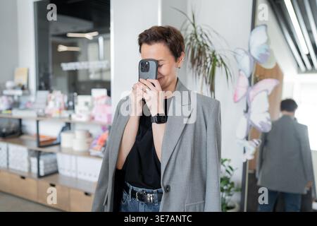 Woman in jacket texting on smartphone and taking call while standing in light modern office. Phone, texting, call, office, woman, business. Stock Photo