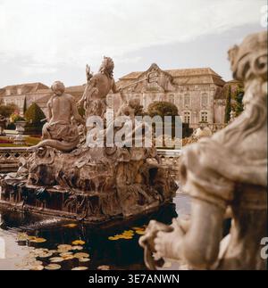 Blick zum Neptunbrunnen, er zeigt Neptun, den römischen Gott des Meeres, auf einem Muschelwagen, umgeben von Tritonen, Nereiden und Delfinen. Die Skulpturen sind allegorisch auf die Herrschaft über Wasser und Natur angelegt – ein Symbol für die Macht und Größe des Kurfürsten im Park von Schloss Schwetzingen. Im Hintergrund sieht man die barocke Schlossfassade. Das Bild von circa 1960, zeigt den Brunnen aus einer ungewöhnlichen Perspektive mit einem stimmungsvollen Lichtspiel und Fokus auf die barocke Pracht der Anlage. Original Dia, 6x6 Stock Photo
