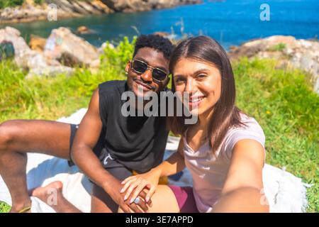 Black boyfriend with caucasian girlfriend taking selfie while having picnic sitting on blanket near seaside on sunny day Stock Photo