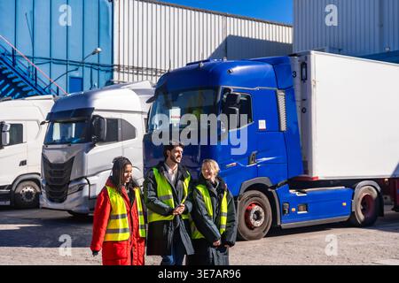Diverse group of engineers wearing hard hats and safety vests walking together near transport trucks at cold storage warehouse on sunny day Stock Photo