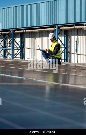Hispanic male engineer with beard wearing hard hat and safety vest using laptop in monitoring performance of large array of solar panels while crou... Stock Photo