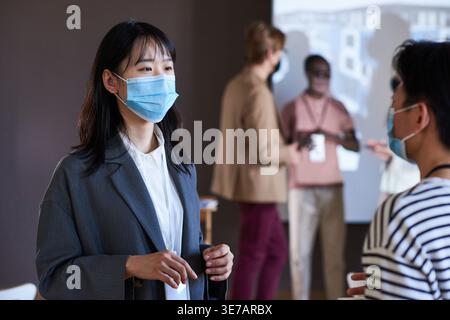 Asian business women in medical masks having conversation while standing in front of their colleagues in conference room during covid Stock Photo