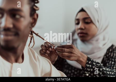 Serious Middle Eastern woman wearing hijab braiding hair of Black Muslim man while standing against white background in studio Stock Photo