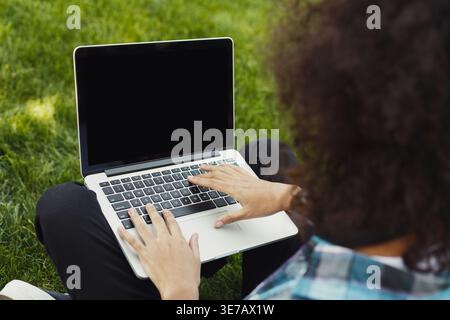 Mixed race woman using laptop while sitting on grass in park on sunny day Stock Photo
